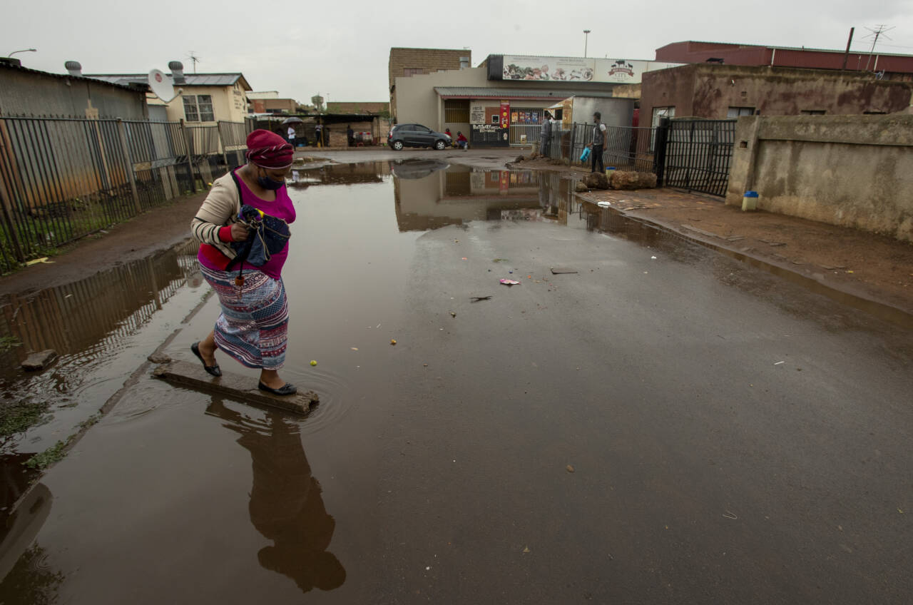 En kvinne med munnbind krysser en oversvømt gate i Thokaza øst for Johannesburg i Sør-Afrika. Landet sliter med et økende antall smittetilfeller og en ny virusmutasjon – i tillegg til utbredt fattigdom og enorme økonomiske ulikheter. Foto: Themba Hadebe / AP / NTB