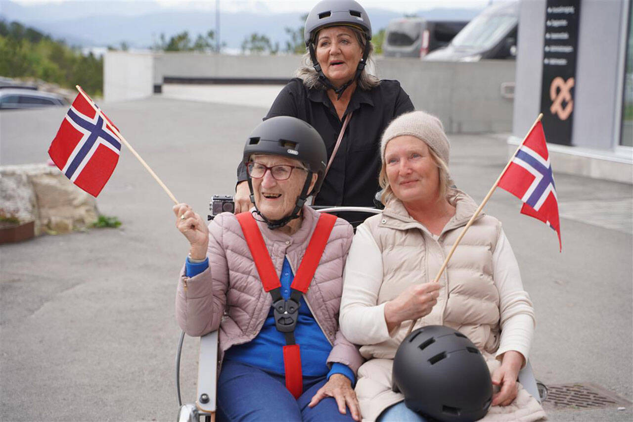 Beboer Birgit (t.v.) sitter sammen med primus motor for prosjektet Randi Rasmussen Bjørlo (t.h.) foran i elsykkeltaxien med hvert sitt flagg. Foto: Kristiansund kommune