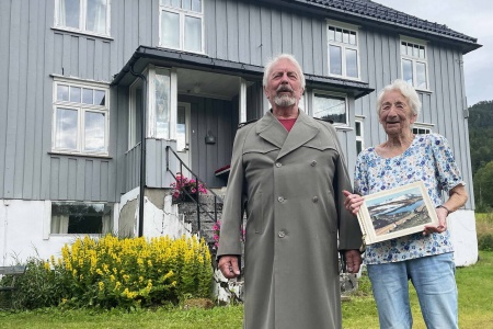 Søskenbarna Geir og Elise Settemsdal framfor «verkstedet» som husa konfeksjonsfabrikk, butikkar og posthus. Geir iført ekte kappe frå fabrikken! Foto: Bernt Bøe